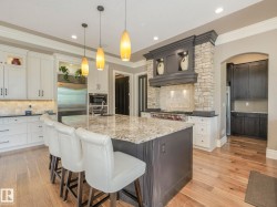 Spacious kitchen featuring a large central island with seating, stone-clad range hood, stainless steel appliances, light-toned cabinetry, and wood-finish flooring - 