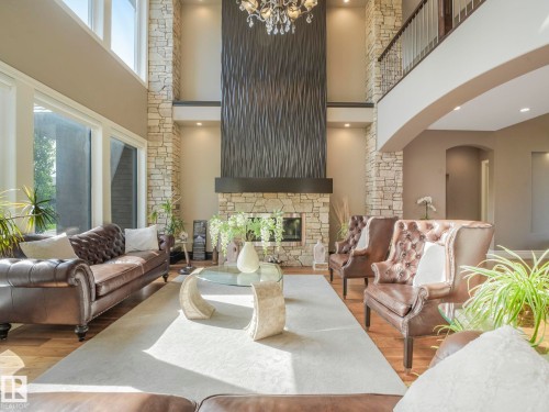 Two-story living area featuring a stone fireplace with a textured dark wood mantle, wood-finish flooring, a neutral-toned area rug, a second-story interior balcony, and a multi-tiered crystal chandelier - 2807 Wheaton Drive, Edmonton, AB 