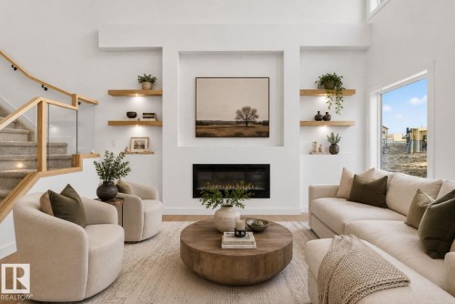Expansive living space featuring a contemporary fireplace with mantel, recessed wall shelving with integrated lighting, wood-finish flooring, and a staircase with glass panel railing and wooden handrail - 105 Wyatt Ridge, Fort Saskatchewan, AB 