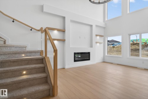 Living area featuring a modern fireplace, high ceilings, large windows, wood-finish flooring, and a carpeted staircase with integrated lighting - 105 Wyatt Ridge, Fort Saskatchewan, AB 