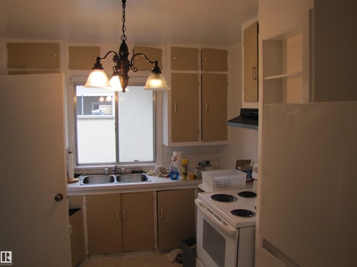 Compact kitchen featuring a double-basin stainless steel sink, cabinetry with various door and shelf configurations, a range hood, and a ceiling-mounted chandelier - 8122 83 Avenue, Edmonton, AB 