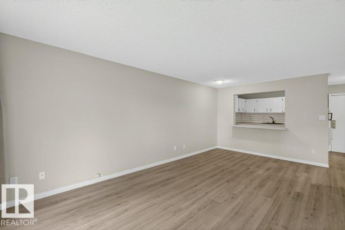 Open-concept living area featuring wood-finish flooring, light gray walls, and a pass-through window to the kitchen - 103 10620 104 Street Nw, Edmonton, AB 