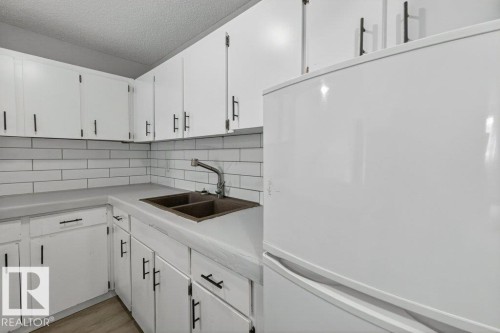 Kitchen featuring white cabinetry with matte black hardware, subway tile backsplash, a double basin sink with gooseneck faucet, light-toned countertops, and a white refrigerator - 103 10620 104 Street Nw, Edmonton, AB 