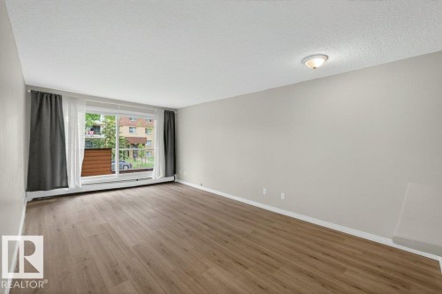 Open living area featuring wood-finish flooring, light neutral wall paint, and a large sliding glass door with a balcony - 103 10620 104 Street Nw, Edmonton, AB 