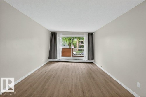 Spacious room featuring light-toned walls, wood-finish flooring, and a large window - 103 10620 104 Street Nw, Edmonton, AB 