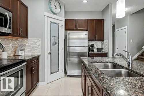 Kitchen featuring dark wood cabinetry, stainless steel appliances, granite countertops, a mosaic tile backsplash, and a pantry with a frosted glass door - 13004 164 Avenue, Edmonton, AB 