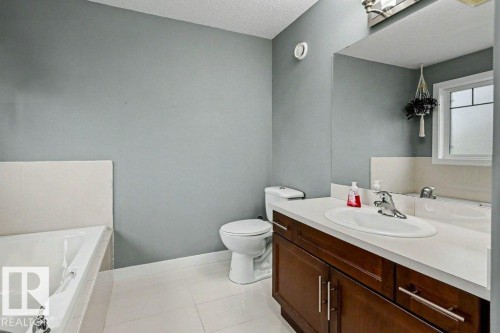 Bathroom featuring a built-in tub with tile surround, a dark wood-finish vanity with an integrated sink, a large wall-mounted mirror, a toilet, and light-toned floor tiles - 13004 164 Avenue, Edmonton, AB 
