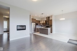 Contemporary open concept living area featuring a built-in electric fireplace, wood-finish flooring, a kitchen island with pendant lighting, stainless steel appliances, and sleek cabinetry - 