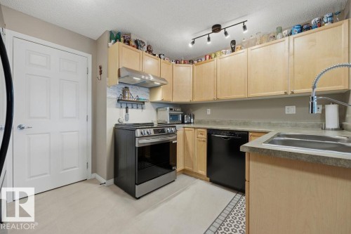 Kitchen featuring light wood cabinetry, stainless steel range with overhead ventilation, black dishwasher, and a double basin sink with a gooseneck faucet - 71 3040 Spence Wynd, Edmonton, AB 