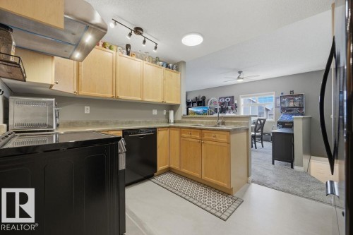 Kitchen featuring light wood cabinetry, a stainless steel range hood, black appliances, and neutral countertops - 71 3040 Spence Wynd, Edmonton, AB 