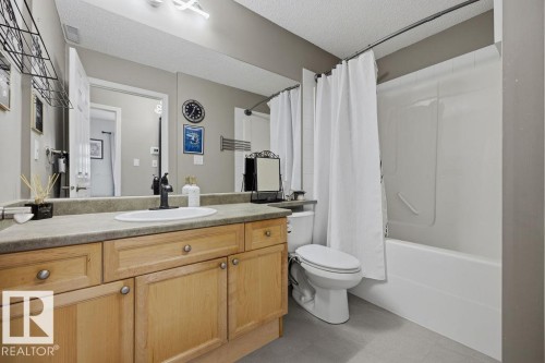 Bathroom featuring a light wood-finish vanity with a single basin sink, a large wall-mounted mirror, a toilet, and a combined bathtub and shower unit - 71 3040 Spence Wynd, Edmonton, AB 