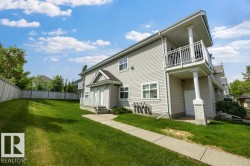 Modern multi-story structure with light-colored siding, featuring a second-story balcony with white railing, a ground-level patio, and a front entry with a small porch - 