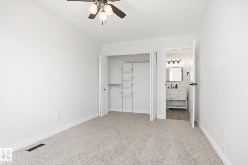 Carpeted room with white walls, a ceiling fan with light fixture, bi-fold closet doors revealing built-in shelving, and a doorway opening to a bathroom with a white vanity and mirror - 857 Erin Place, Edmonton, AB 