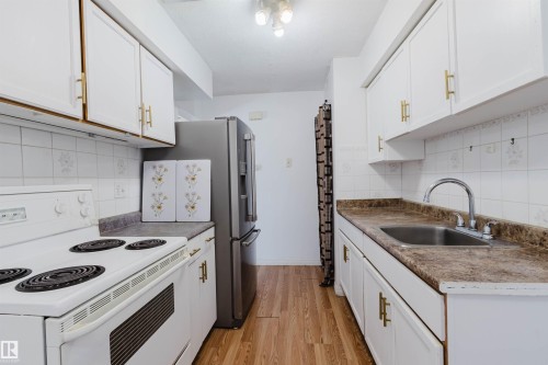 Galley kitchen featuring white cabinetry with gold-tone hardware, wood-finish flooring, white tile backsplash, a stainless steel refrigerator, and a stainless steel sink with a chrome faucet - 4 14310 80 Street, Edmonton, AB 