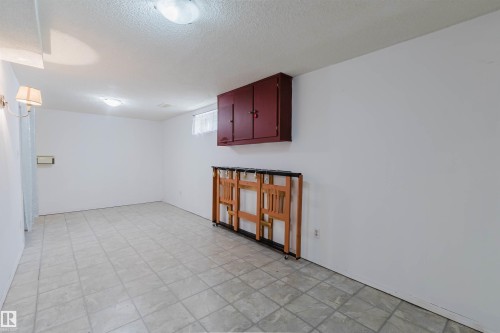 Spacious interior room featuring light-toned square tile flooring, a wall-mounted red wood-finish storage cabinet, a small window with light-colored curtains, and two ceiling-mounted dome lights providing illumination - 4 14310 80 Street, Edmonton, AB 