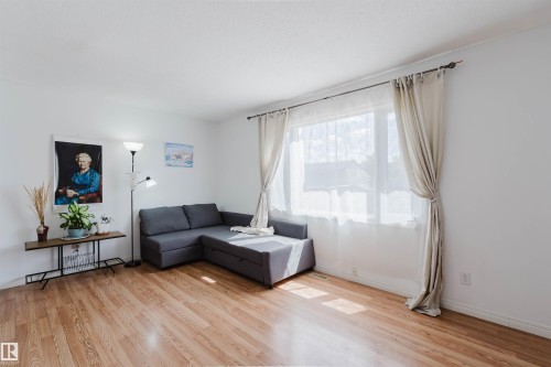 Spacious room featuring wood-finish flooring, white baseboards, and a large window with sheers and drapes - 4 14310 80 Street, Edmonton, AB 
