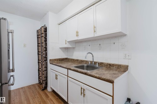 Kitchen featuring white cabinetry with gold-toned hardware, a stainless steel sink with a gooseneck faucet, and white tile backsplash with decorative accents - 4 14310 80 Street, Edmonton, AB 