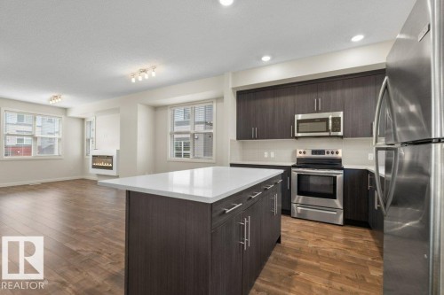 Modern kitchen island with a white countertop and wood-finish cabinetry, stainless steel appliances, recessed lighting, and wood-finish flooring - 125 655 Watt Boulevard, Edmonton, AB 
