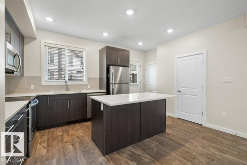 Modern kitchen featuring wood-finish flooring, dark wood-finish cabinetry, stainless steel appliances, a center island with a light-toned countertop, and recessed lighting - 125 655 Watt Boulevard, Edmonton, AB 