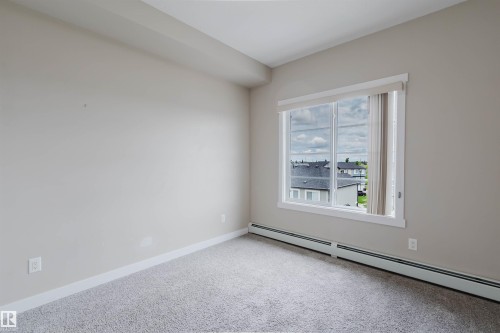 Carpeted interior space featuring a large window with white trim, light-colored walls, and baseboard heating - 402 3670 139 Avenue, Edmonton, AB 