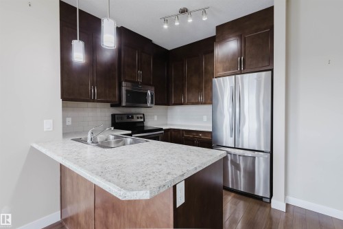 Kitchen featuring dark wood cabinetry, light speckled countertops, stainless steel appliances, and wood-finish flooring - 402 3670 139 Avenue, Edmonton, AB 