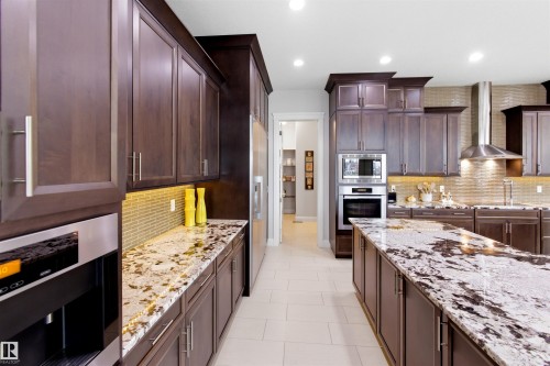 Gourmet kitchen featuring dark wood cabinetry, light tile flooring, stainless steel appliances, a textured tile backsplash, and extensive stone countertops, including a large center island - 9003 16 Avenue, Edmonton, AB 