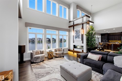 Two-story living area featuring large windows with waterfront exposure, dark wood-finish flooring, a modern linear fireplace with architectural wood accents, and a contemporary kitchen with dark cabinetry - 9003 16 Avenue, Edmonton, AB 