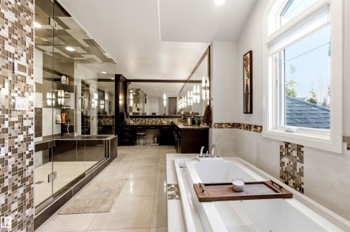Spacious bathroom featuring a large soaking tub, glass-enclosed shower with tiled walls, dual vanities with dark wood cabinetry, and extensive tile detailing - 9003 16 Avenue, Edmonton, AB 