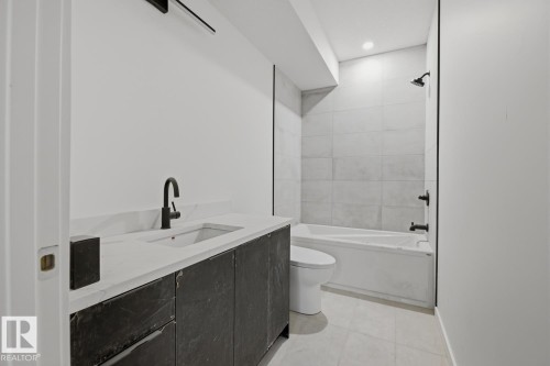 Bathroom featuring a dark wood-finish vanity with a light-toned countertop, a black matte faucet, a built-in bathtub with a shower, and light-toned wall and floor tiling - 11327 122 Street, Edmonton, AB - Indoor Photo Showing Bathroom