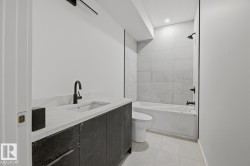 Bathroom featuring a dark wood-finish vanity with a light-toned countertop, a black matte faucet, a built-in bathtub with a shower, and light-toned wall and floor tiling - 