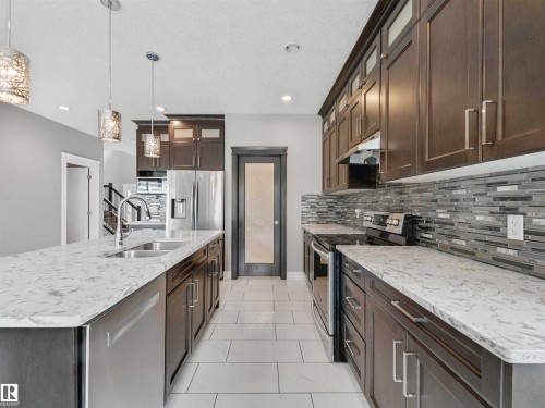 Kitchen featuring an expansive island with dual basin sink and pendant lighting, dark wood-finish cabinetry, light-toned countertops, and a full tile backsplash - 1090 Armitage Crescent, Edmonton, AB 