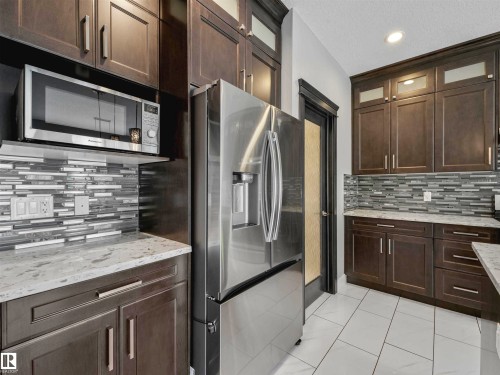 Contemporary kitchen featuring stainless steel appliances, dark wood-finish cabinetry, light-toned countertops, a mosaic tile backsplash, and white tile flooring - 1090 Armitage Crescent, Edmonton, AB 