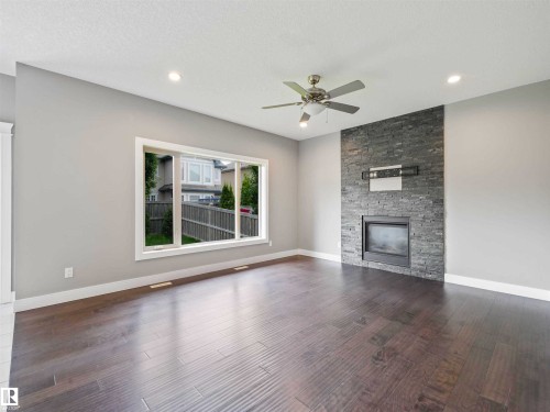 Spacious room featuring wood-finish flooring, a contemporary fireplace with a stacked stone surround, a ceiling fan, recessed lighting, and a large window with white trim - 1090 Armitage Crescent, Edmonton, AB 