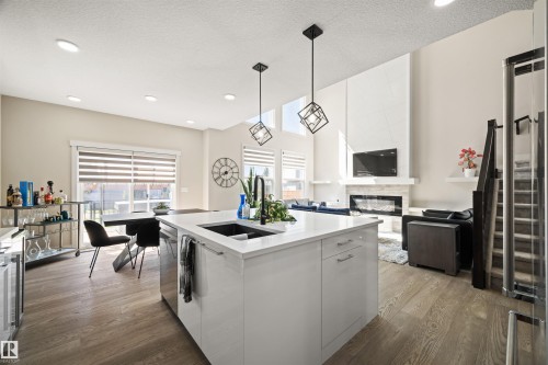 Open-concept living area featuring a large kitchen island with a dark sink, wood-finish flooring, a contemporary fireplace, high ceilings, and modern pendant lighting - 12171 177 Avenue, Edmonton, AB 