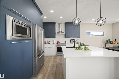 Contemporary kitchen featuring a large central island with a white countertop, two geometric pendant lights, stainless steel appliances, and two-tone cabinetry with blue and white finishes - 12171 177 Avenue, Edmonton, AB 