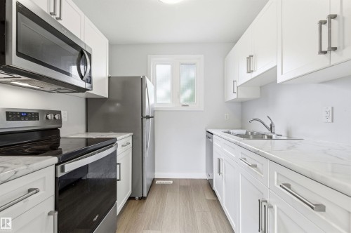 Bright kitchen featuring white cabinetry with brushed nickel hardware, stainless steel appliances, marble-look countertops, and wood-finish flooring - 18353 66 Avenue, Edmonton, AB 