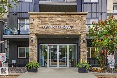 Building entrance featuring stacked stone facade, double glass doors, exterior wall sconces, and planters with greenery - 211 1204 156 Street Nw, Edmonton, AB 