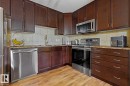 Contemporary kitchen featuring dark wood cabinetry, stainless steel appliances, a tile backsplash, wood-finish flooring, and ample countertop space - 211 1204 156 Street Nw, Edmonton, AB 