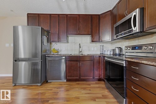 Kitchen featuring wood-finish flooring, dark wood cabinetry, stainless steel appliances, speckled countertops, and a tile backsplash - 211 1204 156 Street Nw, Edmonton, AB 