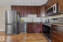 Kitchen featuring wood-finish flooring, dark wood cabinetry, stainless steel appliances, speckled countertops, and a tile backsplash - 211 1204 156 Street Nw, Edmonton, AB 