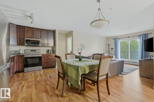 Open-concept living area featuring wood-finish flooring throughout, a modern kitchen with stainless steel appliances, and ample natural light from the balcony access - 211 1204 156 Street Nw, Edmonton, AB 