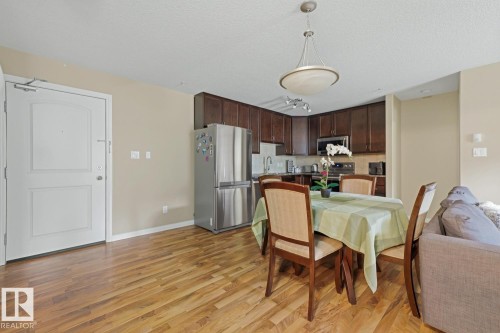 Open-concept living space with wood-finish flooring, a kitchen featuring dark wood cabinetry and stainless steel appliances, and a dedicated dining area with a contemporary chandelier - 211 1204 156 Street Nw, Edmonton, AB 