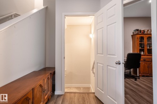 Wood-finish flooring throughout, featuring a white paneled door with a brushed nickel knob, a recessed hallway with wall sconce lighting, and a staircase with a white painted railing - 17562 63A Street, Edmonton, AB 