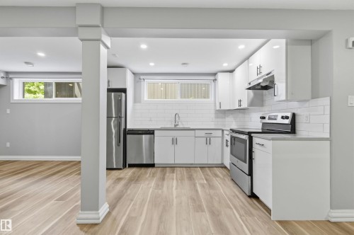 Open-concept kitchen featuring wood-finish flooring, white cabinetry, stainless steel appliances, a subway tile backsplash, and recessed lighting - 7412/7414 83 Avenue, Edmonton, AB 