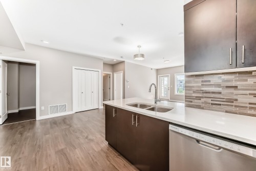 Modern kitchen featuring a double basin stainless steel sink, a stainless steel dishwasher, white countertops, dark wood-finish cabinetry, and a multi-toned horizontal tile backsplash - 303 560 Griesbach Parade Parade, Edmonton, AB 