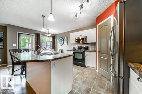 Kitchen featuring a central island with an integrated sink, white cabinetry, dark stone-finish countertops, black appliances, and tile flooring - 8507 2 Avenue, Edmonton, AB - Indoor Photo Showing Kitchen With Double Sink