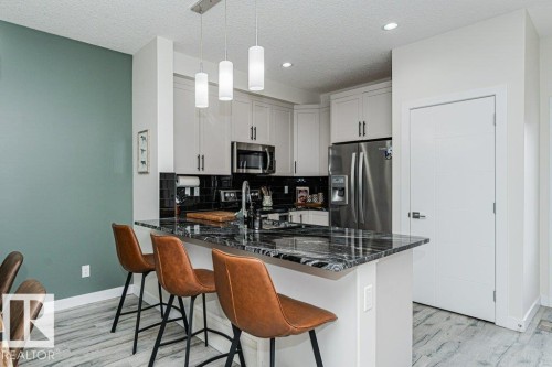 Contemporary kitchen featuring a large island with a dark stone countertop, white cabinetry with dark hardware, stainless steel appliances, black subway tile backsplash, and light wood-finish flooring - 721 Berg Loop, Leduc, AB - Indoor Photo Showing Other Room