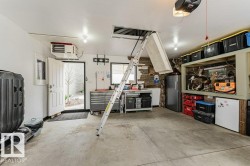Spacious garage featuring a concrete floor, overhead lighting, an attic access ladder, a built-in workbench, and ample storage shelving - 