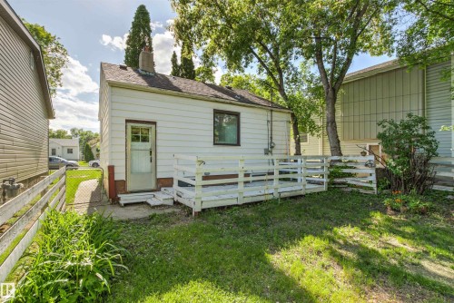 Rear exterior featuring white siding, a small deck, mature trees, and a partial fence - 11417 65 Street, Edmonton, AB 