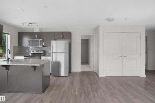 Modern kitchen featuring an island with a built-in sink, stainless steel appliances, dark wood-finish cabinetry, and a subway tile backsplash - 202 142 Ebbers Boulevard, Edmonton, AB 